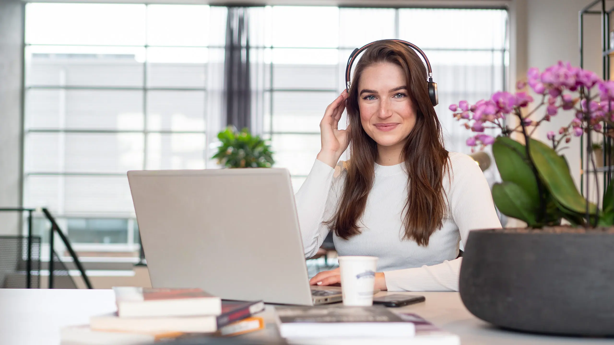 Een vrouw zit achter haar laptop aan een tafel met laptop en headset en belt terwijl ze in de camera kijkt.